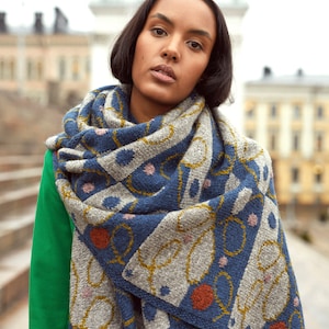 May include: A woman wearing a green jacket with a blue, gray, and yellow patterned scarf. The scarf has a geometric design with small pink and red accents.
