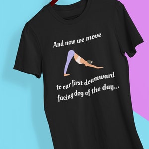 May include: Black t-shirt with white text that reads "And now we move to our first downward facing dog of the day..." and a cartoon illustration of a person doing a downward facing dog yoga pose.