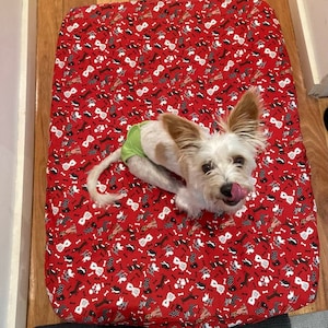 May include: A small white dog wearing a green diaper is lying on a red dog bed with a pattern of black and white dogs and bones.