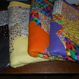 Op de afbeelding: Vier fleecedekens met hondenmotief, gestapeld op elkaar. De bovenste deken is een veelkleurig patchwork met voetafdrukken en de tekst "1 yard hace mailman Potball Cheat Get not marun Sleep Cho dig under fence tleepruni Get news for cookie". De tweede deken is paars met een voetafdrukvorm. De derde deken is oranje met een voetafdrukvorm. De onderste deken is geel met een voetafdrukvorm.