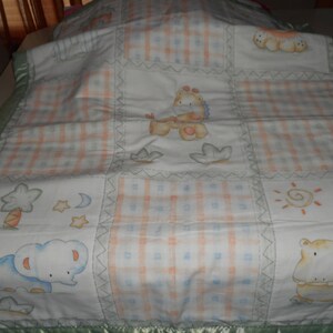 Puede incluir: Una manta de bebé de patchwork con un borde verde. La manta presenta una variedad de diseños de animales, incluyendo un elefante, una jirafa y un hipopótamo. Los diseños están en tonos de azul, amarillo, naranja y verde.