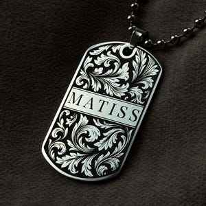Op de afbeelding: Zilverkleurige dogtag-hanger met de naam "MATISS" gegraveerd in een rechthoekige doos. De tag heeft ingewikkeld bloemenkrullenwerk en hangt aan een kogelketting. De achtergrond is een donker, getextureerd oppervlak.