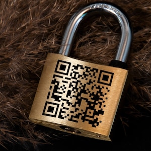 May include: A gold padlock with a black QR code that says "SCAN ME" on the front. The padlock is lying on a brown furry surface.