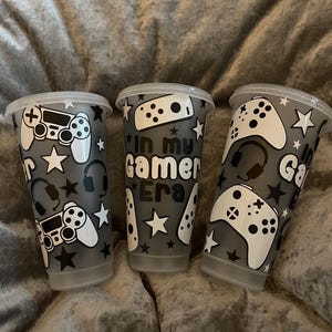 May include: Three clear plastic cups with lids, decorated with white gaming controller graphics, headphones, and stars on a grey background. One cup reads "In my Gamer Era".