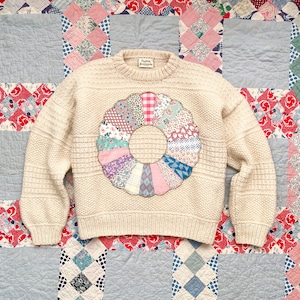 May include: A cream-coloured knit jumper featuring a round, multi-coloured patchwork appliqué on the chest. The appliqué consists of various floral, plaid, and geometric fabric segments arranged in a circular pattern with a scalloped edge.