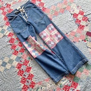 Peut inclure: Pantalon en jean bleu doté d'une taille haute avec un cordon de serrage blanc, deux poches plaquées à l'avant et de grands panneaux rectangulaires en tissu patchwork rose, blanc et bleu cousus sur le devant de chaque jambe, du genou à l'ourlet.