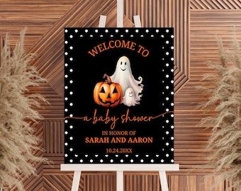 May include: A rectangular black sign with a white polka dot border featuring an orange pumpkin and two white ghosts. The text reads: WELCOME TO a baby shower IN HONOR OF SARAH AND AARON 10.24.20XX. The sign is displayed on a white wooden easel.