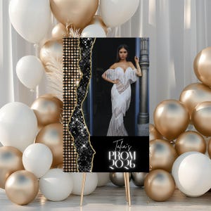 May include: A sign for Talia's Prom 2026, featuring a photo of a woman in a white fringed gown. The sign has a black and gold design with a gold easel. Gold and white balloons are in the background.
