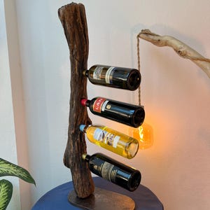 Puede incluir: Un botellero rústico de madera que sostiene cuatro botellas de vino. El botellero está hecho de una rama de árbol desgastada y tiene una base de metal. Las botellas son de varios colores, con etiquetas visibles.