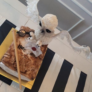 May include: A white owl cake topper with outstretched wings, perched on top of a Harry Potter book cake. The book is decorated with a golden wand and a red wax seal.