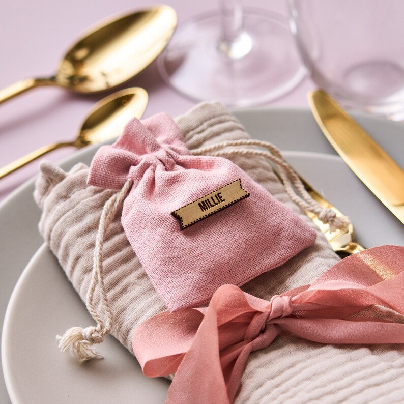 K&ouml;nnte beinhalten: Ein rosa Stoffbeutel mit einem Holzschild, auf dem "Millie" steht. Der Beutel liegt auf einer beigefarbenen Serviette mit einem rosa Band, das darum gebunden ist. Die Serviette liegt auf einem wei&szlig;en Teller mit goldenem Besteck.