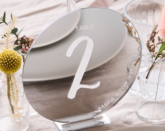 May include: A round, silver, mirrored table number with the number "2" written in white. The table number is on a clear acrylic stand. The text "TABLE" is written above the number and "SASKIA + TOM 12.07.22" is written below the number.