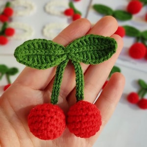 May include: Two red crocheted cherries with green crocheted leaves. The cherries are attached to the leaves by green yarn.