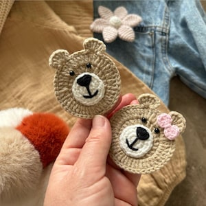 May include: Two crocheted bear face appliques. The bears are tan with white muzzles and black noses and eyes. One bear has a small pink bow. A light pink flower applique and a denim jacket are in the background.