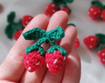May include: A close-up of two crocheted strawberries with green leaves. The strawberries are red with white accents and are held in a hand.