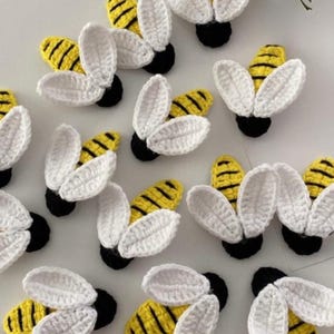 Könnte beinhalten: Gehäkelte Bienen-Applikationen. Jede Biene hat einen gelb-schwarz gestreiften Körper, weiße Flügel und eine schwarze Basis. Diese handgefertigten Applikationen eignen sich für Bastelprojekte und verleihen Kleidung, Accessoires oder Wohnaccessoires eine verspielte Note.