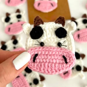 May include: A white, black and pink crocheted cow head appliqué. The cow features a white face, black spots, brown ears and a prominent pink snout with black stitched nostrils. The eyes are small black beads with stitched whiskers.