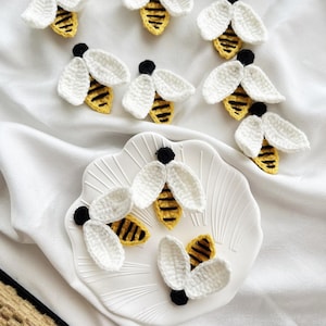 May include: A collection of small, handmade crochet bee appliques featuring white wings, yellow bodies with black horizontal stripes, and black heads. Several of these textile bee embellishments are arranged on a white scalloped plate.