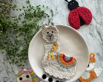 May include: A collection of four crocheted appliques featuring a cream-colored llama with a colorful saddle, a red and black ladybug, a white unicorn with a rainbow mane, and an orange and white fox, all crafted with detailed yarn stitching.