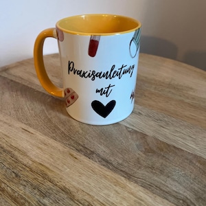 Könnte beinhalten: Weißer Keramikbecher mit gelbem Innenraum und Henkel. Der Becher zeigt den Text "Praxisanleitung mit" in schwarzer Schrift, zusammen mit medizinischen Illustrationen und einem schwarzen Herzen. Der Becher steht auf einer Holzoberfläche.