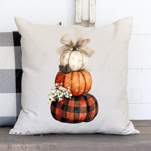 May include: A decorative throw pillow featuring a stack of three pumpkins. The top pumpkin is white with a bow, the middle is orange with white dots, and the bottom is a red and black buffalo plaid. Small pinecones and white flowers are also included.