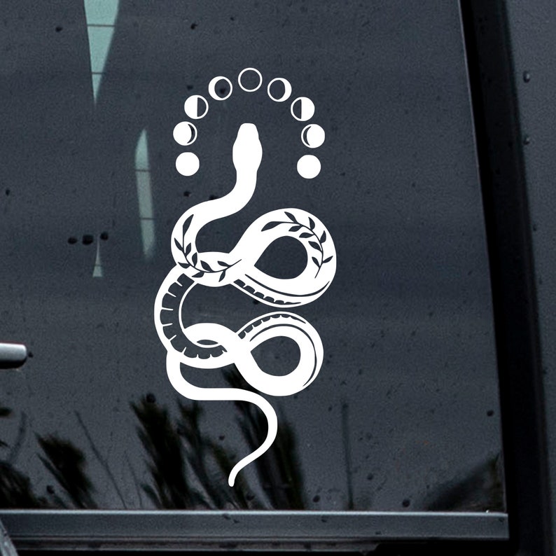 May include: White vinyl sticker of a snake with a crescent moon design above it. The snake is curled in a circle with its tail in its mouth. The crescent moons are arranged in a circle around the snake's head.