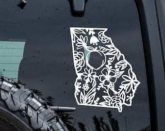 Peut inclure: Un autocollant en vinyle blanc en forme de l'État de Géorgie appliqué sur une vitre de véhicule sombre. Le motif présente un dessin au trait détaillé de diverses fleurs, de feuilles et d'une pêche centrée dans le contour géographique de l'État.