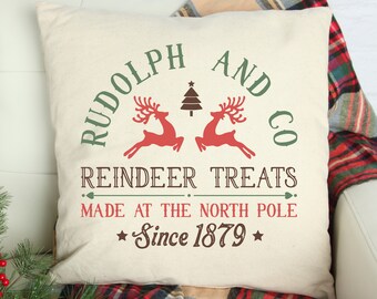May include: A square off-white throw pillow featuring a festive graphic with the text "RUDOLPH AND CO REINDEER TREATS MADE AT THE NORTH POLE Since 1879". The design includes two red reindeer silhouettes, a small brown Christmas tree, and green text.