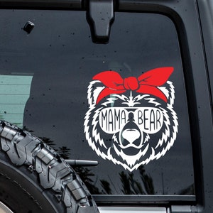 May include: White vinyl decal of a bear wearing a red bandana and sunglasses with the text "Mama Bear" on the sunglasses. The decal is on a black car window.