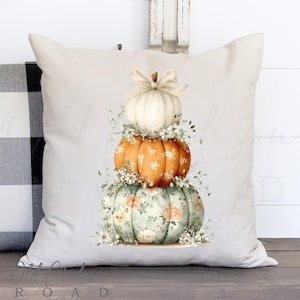 May include: A decorative pillow featuring a stack of three pumpkins. The top pumpkin is white with a bow, the middle is orange with white dots, and the bottom is green with a floral pattern. White flowers and greenery surround each pumpkin.