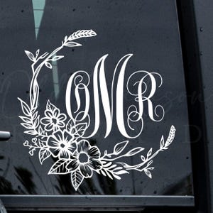May include: A white vinyl decal featuring a decorative floral wreath design that curves around the letters OMR in a stylized script font. The graphic includes detailed leaves, stems, and flower blossoms arranged in a semi-circular pattern.