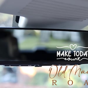 以下が含まれることがあります： 黒い車のバックミラー。白い文字で「MAKE TODAY count」と書かれ、その上に茶色の筆記体で「Old Madison Road」と書かれています。テキストはデカールまたはステッカーです。