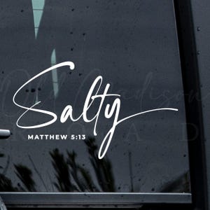 Può includere: Adesivo bianco con la parola "Salty" in corsivo, con "Matthew 5:13" sotto. L'adesivo è applicato su una superficie scura, probabilmente un finestrino dell'auto. Il design è semplice ed elegante.