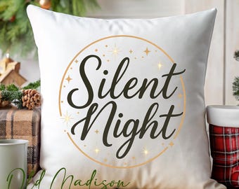 May include: White square pillow with the words "Silent Night" in a black script font, encircled by a gold ring with star accents. The pillow is on a wooden surface, with Christmas decorations in the background.
