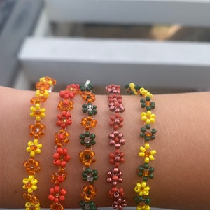 May include: Five beaded bracelets with flower designs in shades of yellow, orange, red, and pink. The bracelets are worn on a wrist, showcasing the intricate beadwork and color variations. Each bracelet features a different color scheme.