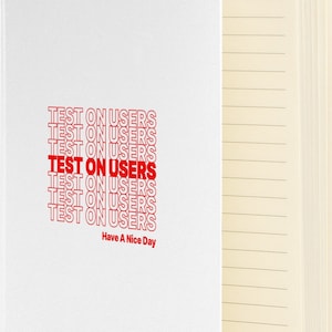 Op de afbeelding: Een wit notitieboek met een rode en witte afbeelding waarop meerdere keren "TEST ON USERS" staat. Het notitieboek heeft gelinieerde pagina's en een witte omslag.