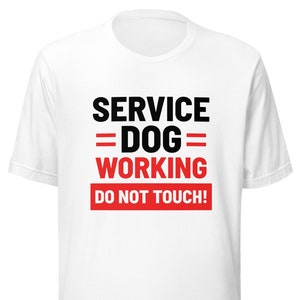 Op de afbeelding: Wit t-shirt met de tekst "SERVICE DOG WORKING DO NOT TOUCH!" in zwart en rood. De tekst staat in het midden op de voorkant van het shirt. Het shirt heeft korte mouwen en een ronde hals.