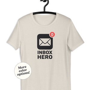 May include: A light beige t-shirt featuring a black envelope graphic with a red notification bubble displaying "0" and the words "INBOX HERO". A small white circle with the text "More color options!" is in the lower left.
