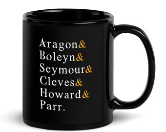 May include: A black ceramic mug with a curved handle. The mug features white text with gold accents, listing names: Aragon, Boleyn, Seymour, Cleves, Howard, and Parr. The mug is designed for beverages.