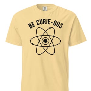 Puede incluir: Una camiseta amarilla con un gráfico de átomo negro y el texto "BE CURIE-OUS".