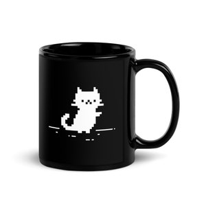 Puede incluir: Una taza de café de cerámica negra con un diseño de gato pixelado blanco. El gato está mirando hacia adelante con la cola enroscada detrás de él.