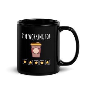 Pode incluir: Caneca de café de cerâmica preta com o texto "I'M WORKING FOR" acima de um gráfico de xícara de café e cinco ícones de classificação por estrelas. O gráfico da xícara de café é marrom com uma tampa amarela e um design circular. As estrelas são amarelas.