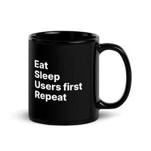 May include: Black ceramic coffee mug with white text that reads "Eat Sleep Users first Repeat".