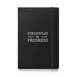 May include: Black leather-bound journal with a white inscription that reads "SCREENPLAY in PROGRESS" with a small floral design.