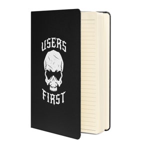 Puede incluir: Cuaderno negro con las palabras "USERS FIRST" y un gráfico de calavera en blanco. El cuaderno está abierto, revelando páginas rayadas. La cubierta tiene un acabado mate.