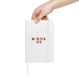 May include: White notebook with a black elastic closure and the text "AUF DEUTSCH, BITTE!" in yellow, red, and black.