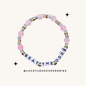 May include: Pink and white beaded bracelet with gold accents and the words "Beat the Odds" spelled out in white letter beads.