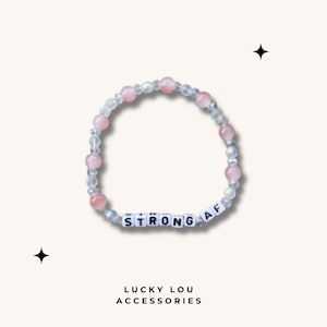 May include: A pink and white beaded bracelet with the words "STRONG AF" spelled out in white letters.