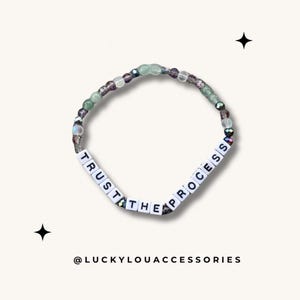 May include: A beaded bracelet with the words "TRUST THE PROCESS" spelled out in white letters. The beads are a mix of iridescent green, purple, and clear.