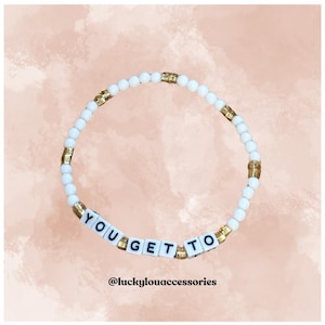 May include: White beaded bracelet with gold accents spelling out "YOU GET TO" in black lettering. The bracelet is on a pink background. @luckyyouaccessories